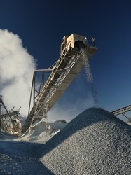 Fine crushed stone is piled up from the conveyor belt of stone crushing equipment at a mining plant against a background of stone dust and a blue sky, close-up. Mining equipment.