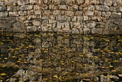 Closeup of stone wall and its reflection in pond of still water with autumn leaves floating on surface.
