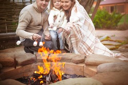 Cheerful young family with little daughter toasting marshmallow over bonfire while enjoying time together in patio of country house
