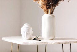 Small marble table with decorations, white vase with dry flowers, little buddha head statue and burning incense stick on stand in modern simplistic apartment environment