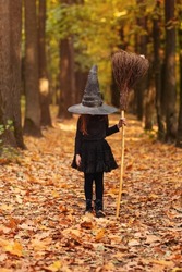 Cute happy little redhaired girl dressed in witch costume standing with broom over autumn forest background with copy space, holding her hat with hands. Holiday of halloween celebration concept