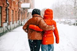 Back view of anonymous women in outerwear hugging each other and walking on snowy city street on winter day