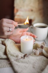 Female hands with lit match lighting burning candle on windowsill for calm and coziness at home, woman trying to create cozy warm and intimate atmosphere in cold season. Hygge lifestyle concept