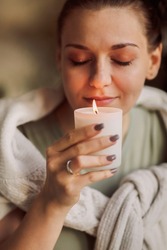 Side portrait of young calm woman holding lit candle in hands, closing eyes bringing close to her face and inhaling its aroma, smelling familiar scent while enjoying happy moment of comfort