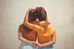 Strong female friendship. Rear view two teen girls best friends holding hands behind back and hugging while standing in front of beige wall outdoors