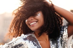 Portrait of overjoyed glamour african american woman with glitter on face in silver sequin dress enjoying outdoor party or event, mixed race female in festive wear with eyes closed feeling happy