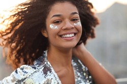 Close up portrait of overjoyed charming african american woman with curly hair and glitter on cheekbones smiling happily at camera while moving dancing outdoors, festive atmosphere in the air