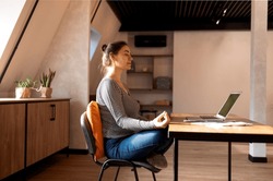 Stress relief during freelance work. Brunette woman freelancer sitting in lotus pose and meditating with hands in mudra gesture on chair in front of open laptop on table, working from home