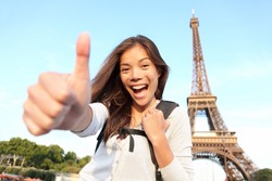 Paris Eiffel tower tourist happy backpacking in Europe. Cheerful smiling woman tourist showing thumbs up success sign in front of Eiffel Tower, Paris. Beautiful Asian Caucasian female model.
