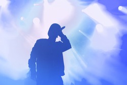 Stock photo of young rap singer with mic in hand singing popular song on stage in blue lights. Hip hop artist performing live on scene in music hall. Rapper with microphone in royalty free backgrounds