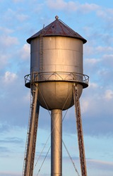 An old metal water tank, in the light of the morning sun, stands tall against a cloudy blue sky in America's Midwest.