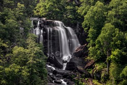The top section of Upper Whitewater Falls, a waterfall near Cashiers in western North Carolina, cascades and plunges over rocks in the mountain forest on a sunny day.