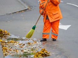 Worker cleaning street with broom tool. Street sweeper with orange jacket cleaning road from fallen leaves in autumn. Janitor cleaner sweep leaves. Municipal worker with broomstick, cleaning service