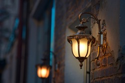 Lantern with yellow warm light on facade of building. Twilight on city street, building illumination. Retro lantern lighting, warm light glow. Street lights, illumination and vintage lantern