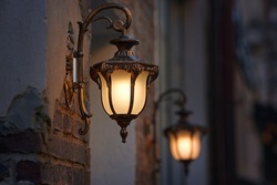 Old street lantern lighting on facade of building. Twilight on city street, building illumination. Retro lantern lighting, warm light glow. Street lights, illumination and vintage lantern.