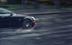 Car driving fast through puddle at heavy rain, dangers of aquaplaning, water splashing over the car. Car driving at heavy rain in evening. Dangerous driving conditions. MOTION BLUR