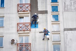  Industrial alpinist at height on rope, plastering wall with trowel. Industrial climber repairing house facade. Rope access job, construction workers repair and restore facade of high building.