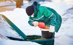 Municipal worker in uniform scoop sand from grit bin, spreading deicing chemicals on slippery sidewalk. Sanding street, prevent injury and slipping accident