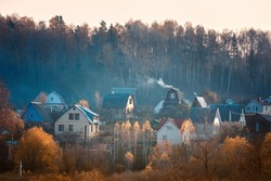 Rural landscape, buildings with smoke from chimney on autumn foggy sunset. Village with houses, smoke rising from chimney of house.. Cottages surrounded by forest, small rural village