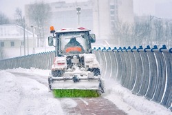 Tractor sweep snow with rotating brush and snowplow from pedestrian zone on bridge, sidewalk snow management during blizzard. Snow plow service remove snow and ice from road, winter road maintenance