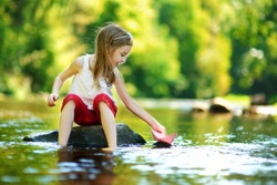Cute little girl playing with paper boat by a river on warm and sunny summer day