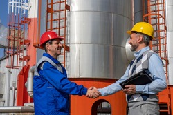 Worker in Uniform and Businessperson Shaking Hands Against Power Plant or Oil Refinery Storage Tanks. Worker in Personal Protective Equipment Meeting with Businessperson. Teamwork Concept.