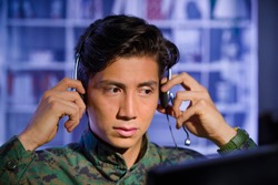 Portrait of handsome soldier wearing a military uniform, watching at his computer and fixing his headphones around the head, ready to give an advice, in a blurred background