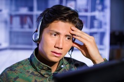 Portrait of worried soldier wearing a military uniform, touching his head and watching at his computer with his headphones around the hea, ready to give an advice, in a blurred background