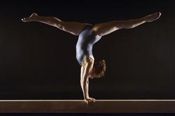 Side view of a female gymnast doing split handstand on balance beam against black background