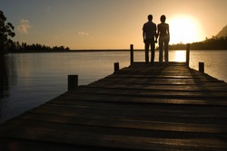 Couple standing on dock by lake holding hands back view.