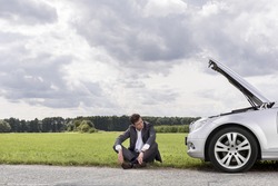 Full length of worried businessman sitting by broken down car at countryside