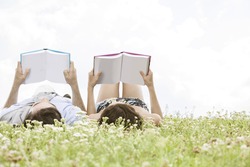 Relaxed young couple reading books while lying on grass against sky
