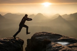 Strong mountain climber hiking and jumping over  the summit ridge of a peaks at sunset. Man takes leap of faith off of rock outcropping