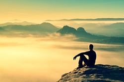 Hiker in black on the rocky peak. Wonderful daybreak in mountains, heavy mist in deep valley. Man sit on the rock.