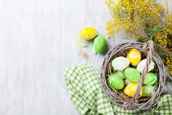 Colorful easter eggs in basket and mimosa flowers on wooden table.  Top view with copy space 