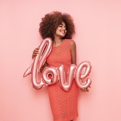 A beautiful young African-American woman with a balloon in the shape of the inscription love on a pink background. Woman on Valentine's Day. Symbol of love