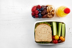 Healthy lunch boxes with sandwich, fresh vegetables, fruits and nuts on white wooden background. From top view