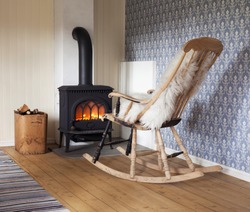 Scandinavian interior: wood burning stove, box of firewood and old restored rocking chair in living room corner. Rag carpet on oiled wooden floor. White sheep skin in rocking chair. 