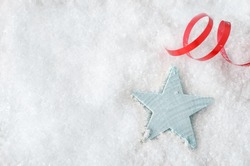 Overhead Christmas background shot.  A pale blue wooden star, resting in white artificial snow, with red swirl of ribbon above and copy space to the left.