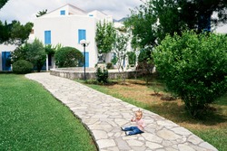 Little girl sits on a cobbled path in the garden in front of the house