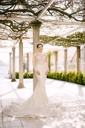 Bride in a mermaid dress stands near a column in an old patio entwined with a vine