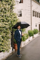 Groom stands under an umbrella near a building entwined with blooming greenery