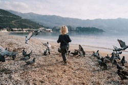 Little girl walks along the seashore among a flock of pigeons. Back view