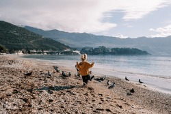 Little girl walks along a pebble beach among pigeons. Back view