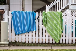 Two striped beach towels and a hat or garment hung over white picket fence by sidewalk in a beach town in South Carolina, USA, for motifs of vacation, recreation, air circulation. Foreground focus.
