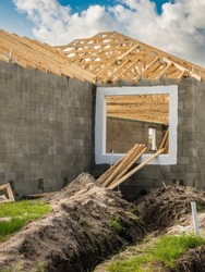 Trench for conduit in front of window opening of single-family house under construction, with wooden roof trusses on concrete shell, in a suburban development in southwest Florida