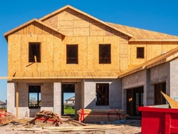 Front exterior of single-family house under construction, with stacks of lumber and a dumpster (bottom right) near two-car garage, in a suburban development on a sunny morning in southwest Florida