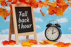 Daylight Saving Time fall back sign with alarm clock and standing blackboard on weathered wood with fall leaves