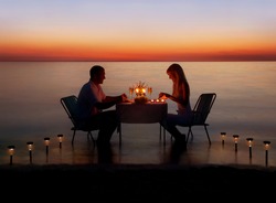 Loving couple share a romantic dinner with candles and lanterns light way at sea beach in water against wonderful sunset