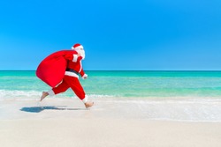 Christmas Santa Claus running with big sack full of gifts hurry on present for children along ocean tropical sandy beach - xmas travel vacation discounts and travel agencies price reductions concept